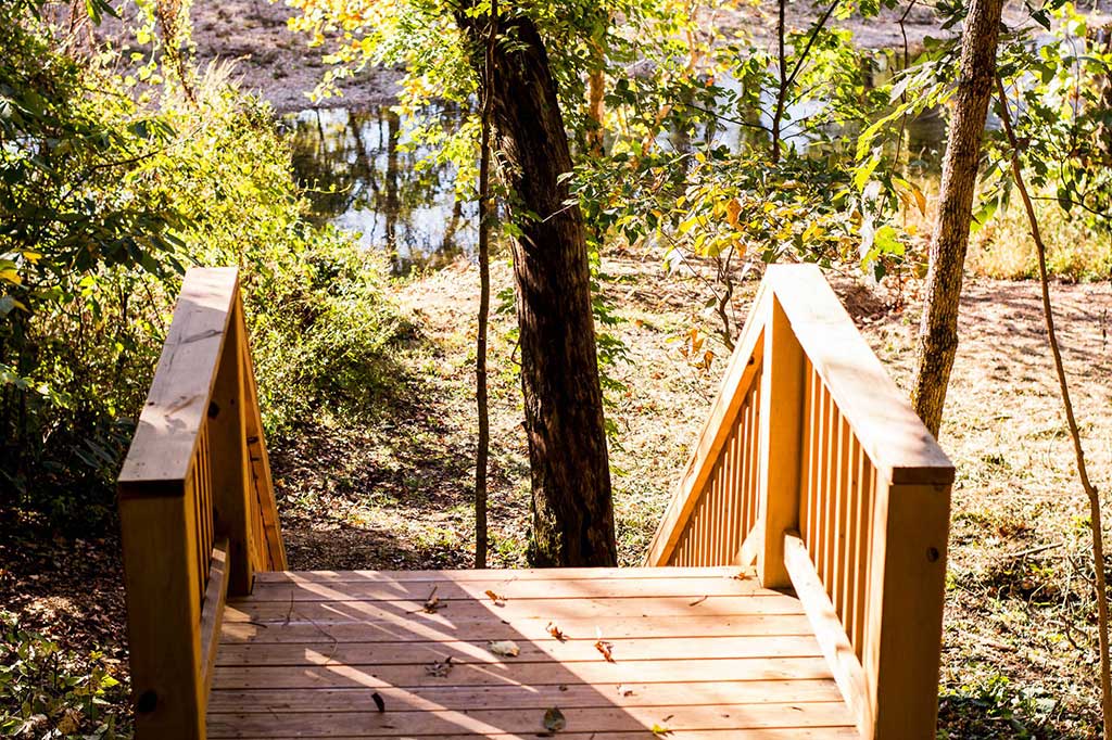 A wooden footbridge with railings leads down toward a forested area with a small pond or creek in the background. Sunlight filters through the trees, casting shadows on the bridge.