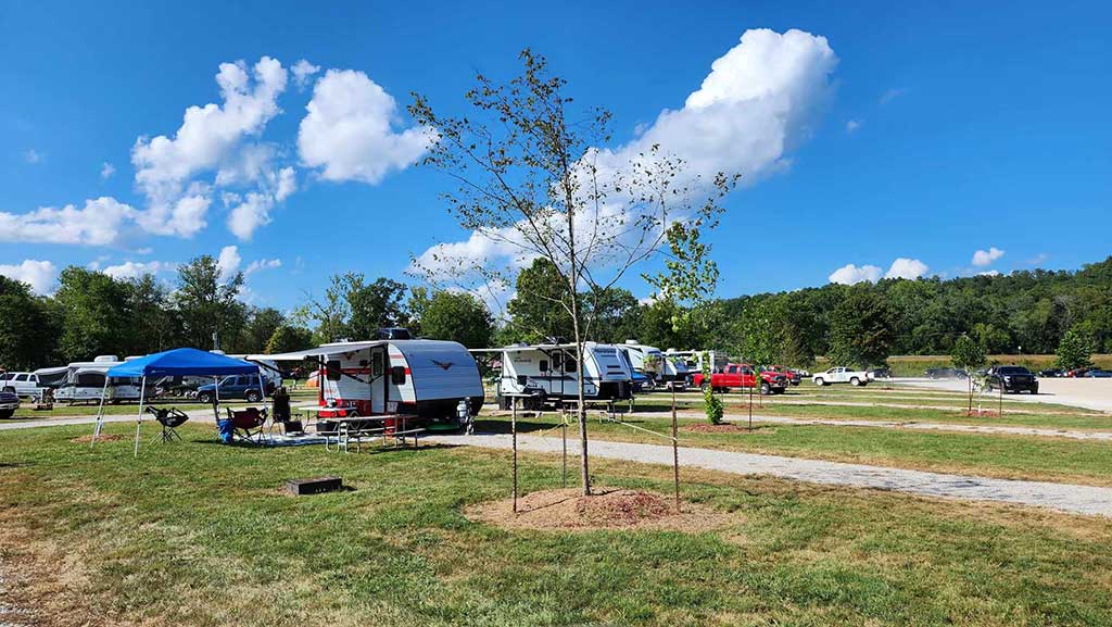 A grassy campground with several RVs and trailers parked on paved areas, some with awnings and outdoor furniture; a few cars and trucks are also visible under a blue sky with fluffy white clouds.