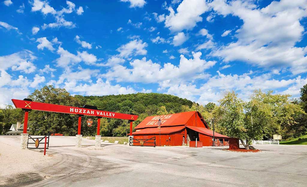 A bright red barn-like building with "Huzzah Valley" written on it sits under a blue sky with scattered clouds, surrounded by trees and greenery in a rural setting.