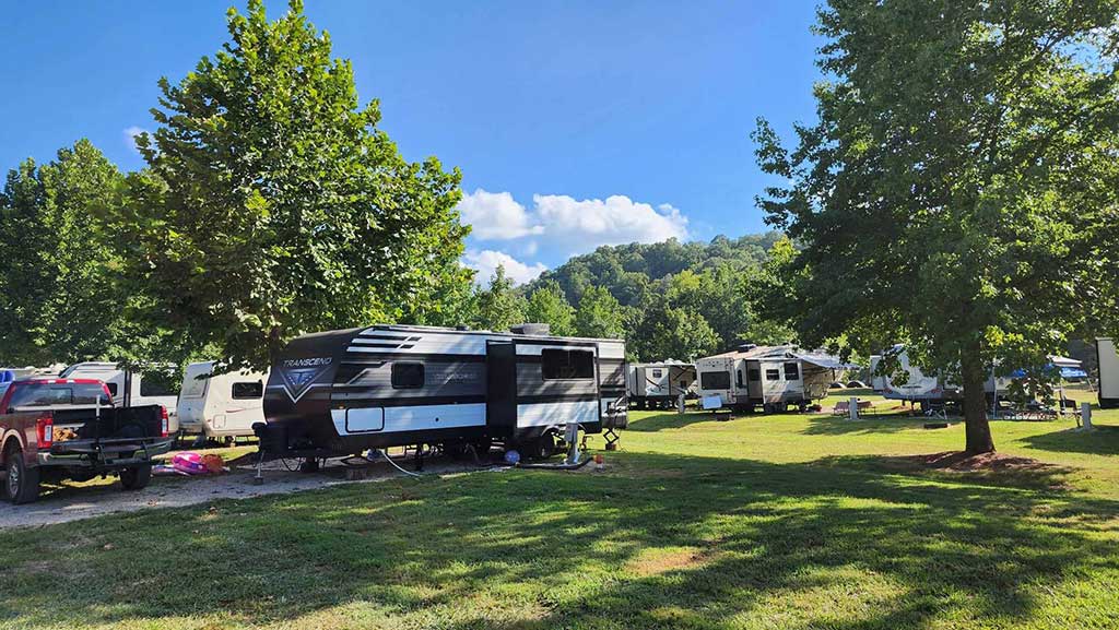Several RVs and campers are parked on a grassy area surrounded by trees under a clear blue sky, with hills and fluffy clouds in the background.