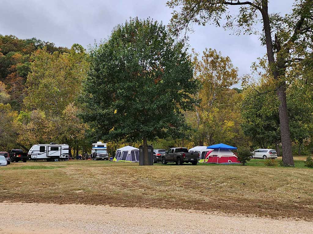 A campground with parked cars, RVs, and several tents set up on grassy areas surrounded by trees with green and yellow foliage under a cloudy sky.