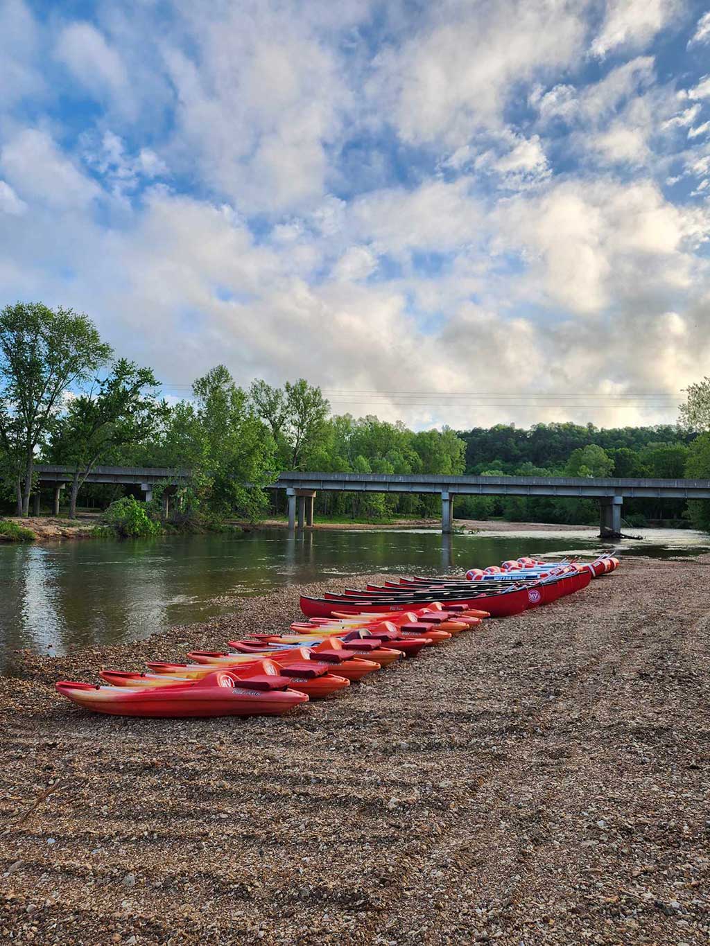 A row of red and orange kayaks lined up on a rocky shore beside a calm river, with a bridge and green trees in the background under a partly cloudy sky.
