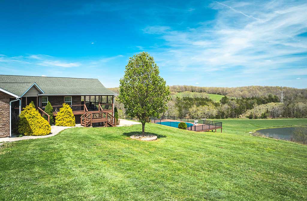 A house with a porch sits on a grassy hill with a tree in the yard, a fenced pool nearby, and rolling green hills and a pond in the background under a bright blue sky.