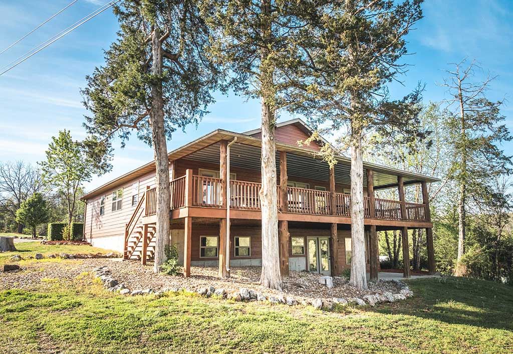 A two-story wooden house with a large wraparound porch, elevated on stilts, surrounded by tall trees and a grassy yard under a clear blue sky.