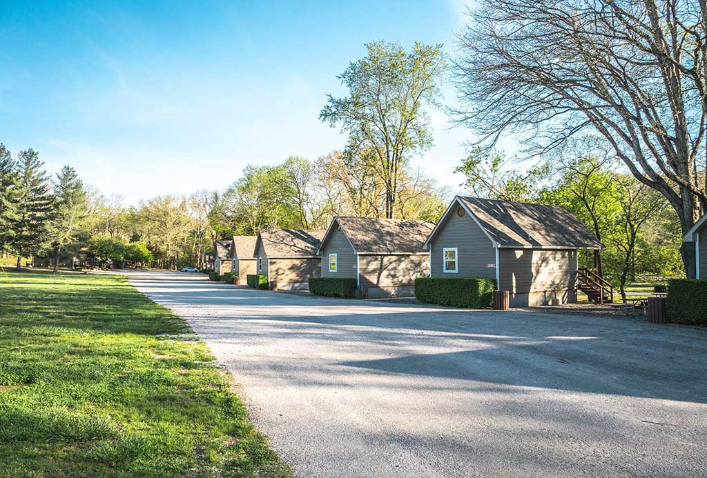 A row of small gray cabins lines a gravel road, surrounded by green grass and tall trees under a clear, sunny sky.