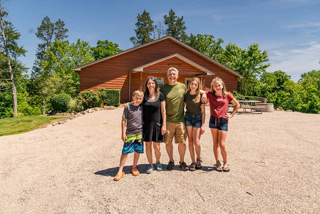 A smiling family of five stands together outside on a gravel driveway in front of a wooden house, surrounded by green trees on a sunny day.
