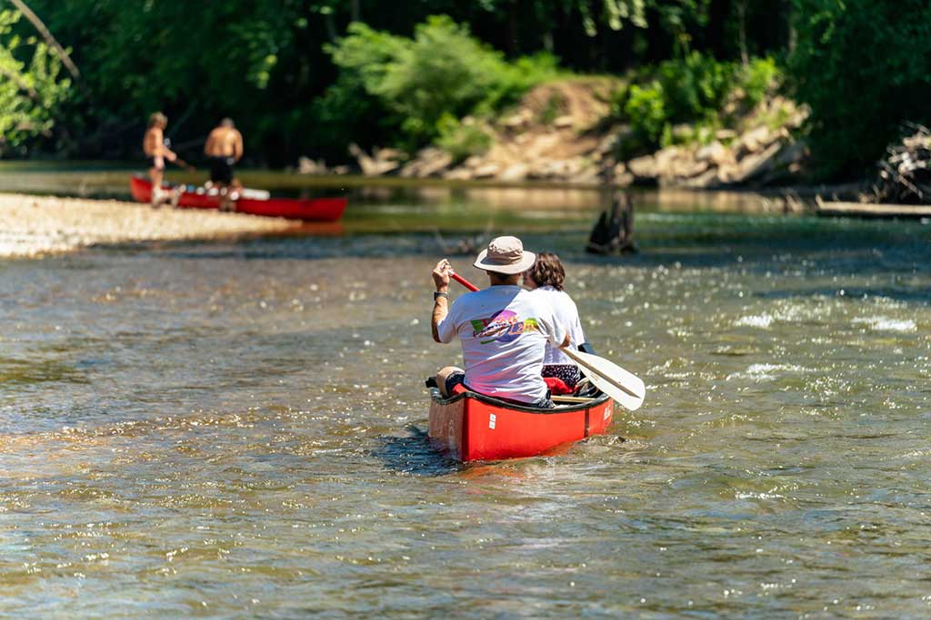 Two people paddle a red canoe down a shallow, sunlit river. In the background, two other people stand near another red canoe by the riverbank, with trees and greenery surrounding the scene.