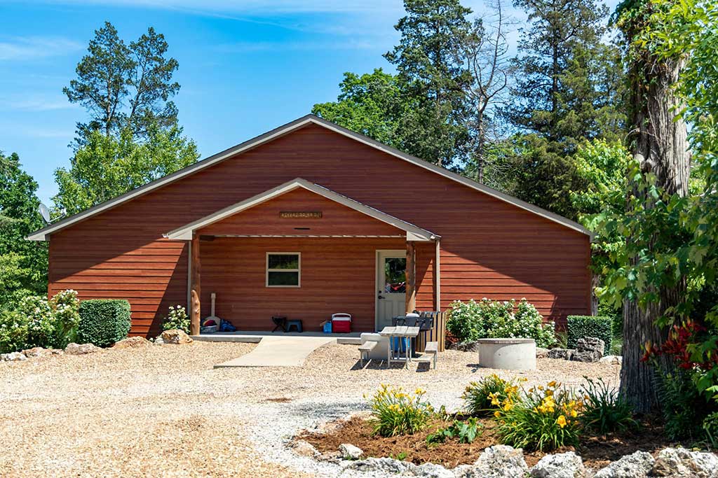 A small red wooden cabin with a covered porch sits among trees and shrubs. Outdoor chairs and various items sit near the entrance, and yellow flowers bloom along a gravel path under a clear blue sky.