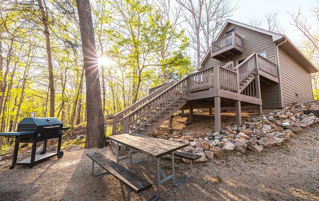 A wooden cabin on a rocky slope with a large staircase, surrounded by trees. In front, there is a picnic table and a barbecue grill, with sunlight streaming through the forest.