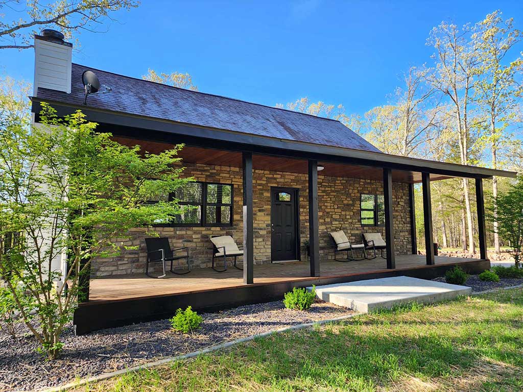 A cozy stone cabin with a dark front porch and wooden ceiling, surrounded by trees and greenery, sits under a clear blue sky. Three chairs and a bench are arranged on the porch, providing a welcoming outdoor space.