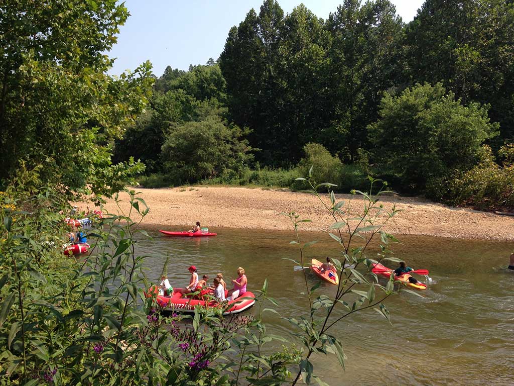 People float down a river in inflatable rafts and kayaks near a sandy riverbank, surrounded by dense green trees and foliage on a sunny day.