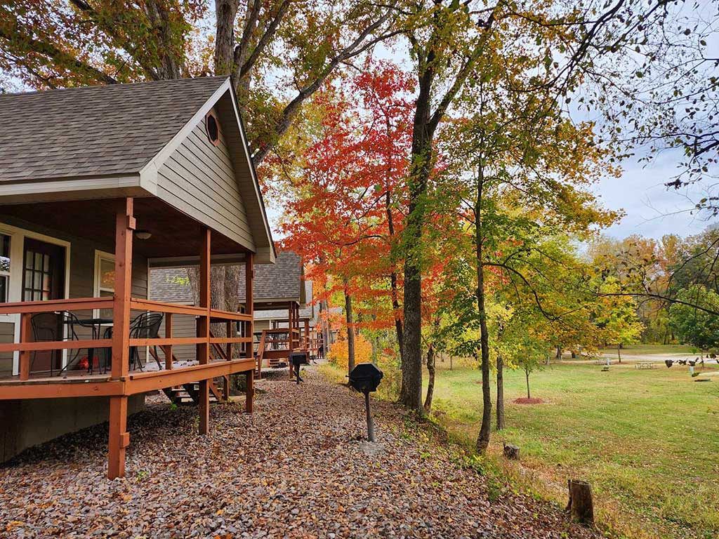 Cabins with covered porches sit among trees with autumn foliage. Fallen leaves cover the ground, and a black barbecue grill stands between the cabins and the grassy open area.