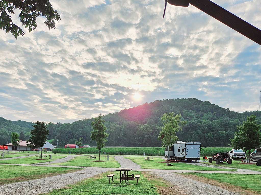 A scenic campground at sunrise with scattered clouds, picnic tables, a few RVs, green grass, trees, gravel paths, and forested hills in the background.