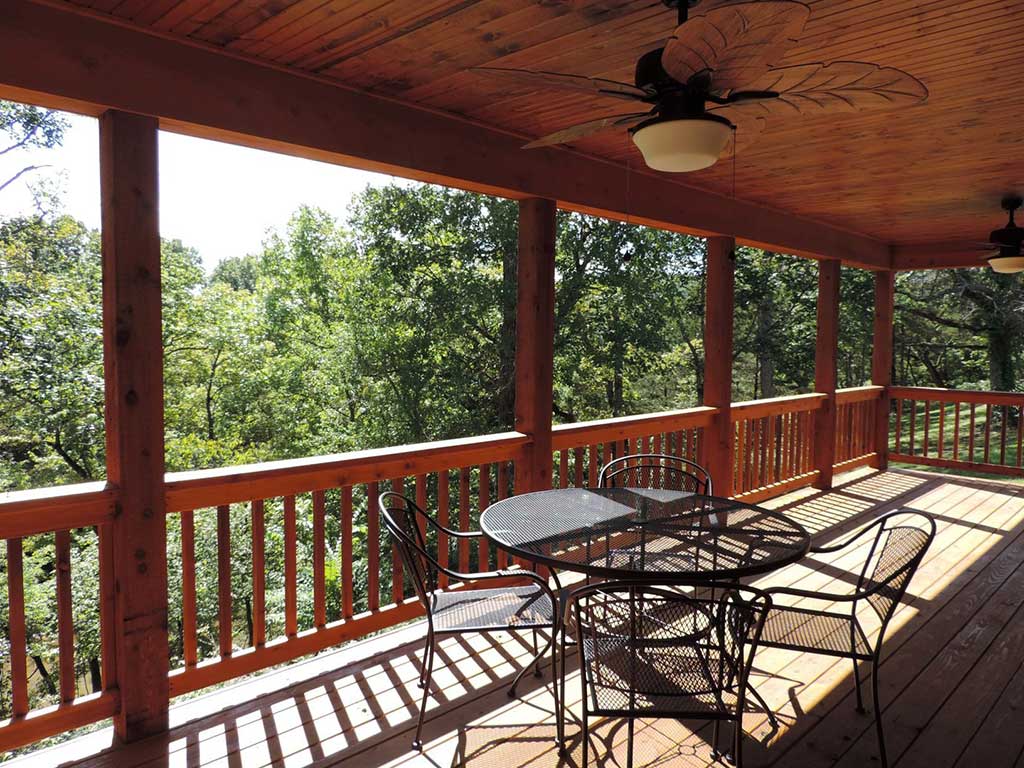 A wooden porch with a ceiling fan, metal table, and four chairs overlooks a lush, green forest. Sunlight filters through the trees and casts shadows on the deck’s floor.