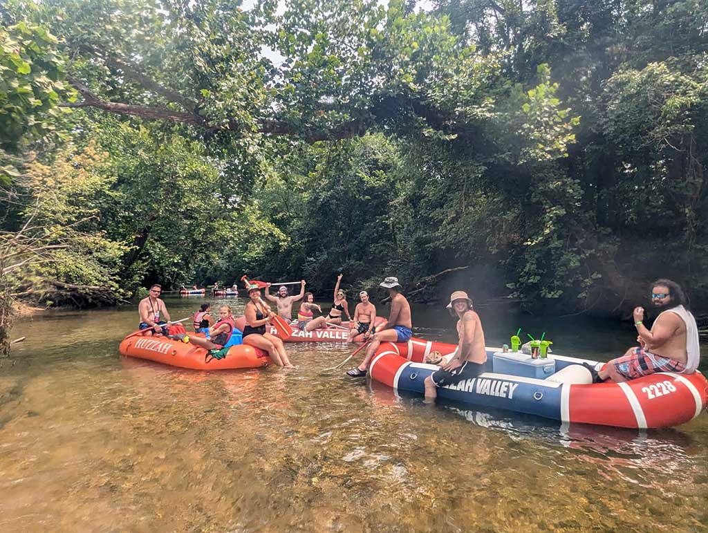 A group of people relax on inflatable rafts in a shallow, clear river surrounded by trees. They are wearing swimsuits and sunglasses, enjoying the sunny day and socializing.
