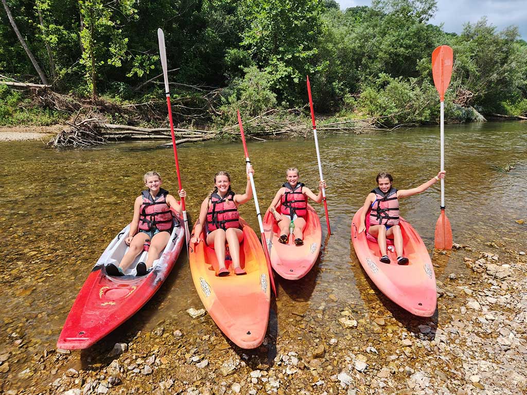 Four children wearing life jackets sit in red kayaks on a shallow, rocky riverbank, each holding a paddle upright. Lush green trees and a fallen log line the river in the background.