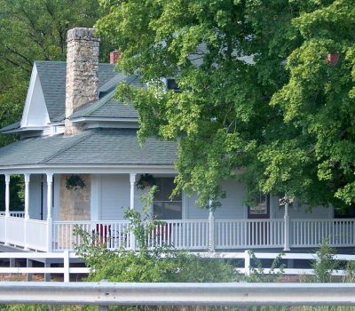 A light blue house with a large wraparound porch, stone chimney, and green roof is partly obscured by leafy trees. A white railing and hanging plants decorate the porch. A metal guardrail runs along the front.