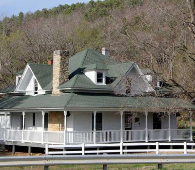 A white, two-story house with a green roof, stone chimneys, and a wraparound porch sits among leafless trees near a wooded hillside. A white fence and guardrail run along the front.