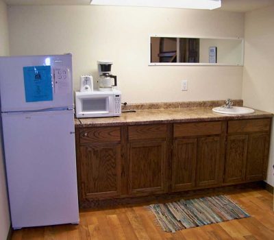 A small kitchenette with a white refrigerator, microwave, and coffee maker on a brown countertop, wooden cabinets below, a sink, a trash can, and a rug on the wooden floor. A mirror hangs above the counter.