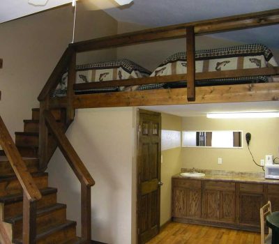 A small room with a wooden loft holding two beds, accessible by stairs. Below, there’s a kitchenette with cabinets, a sink, microwave, coffee maker, and a mirror above the counter.