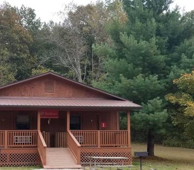 A small wooden cabin with a front porch stands among green trees and grass. The cabin has two doors labeled 1 and 2, steps leading up, and two barbecue grills in the grassy yard.