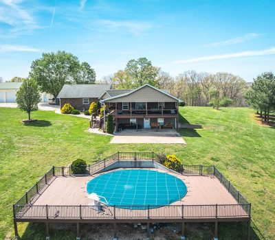 A house with a porch sits on a large grassy lot, with a fenced, covered circular pool in the foreground and a detached garage to the left under a clear blue sky.