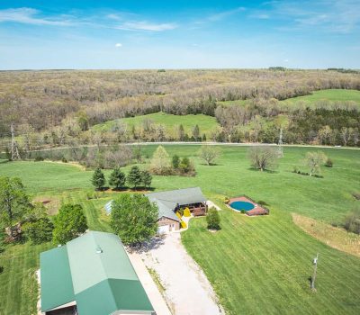 Aerial view of a rural property with a house, barn, round swimming pool, trees, open grassy fields, and a pond, surrounded by dense forest under a blue sky.