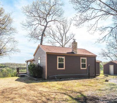 A small brown cabin with a chimney sits among leafless trees, overlooking a distant green landscape. A small shed is nearby, and the ground is covered with gravel and sparse grass under a bright blue sky.
