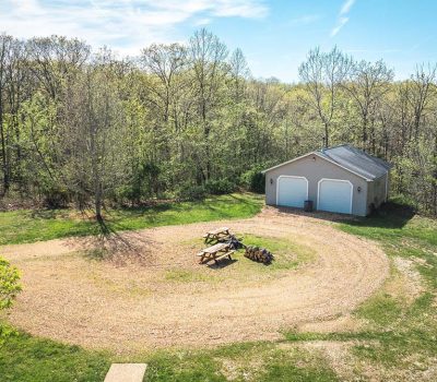 A gravel driveway curves around a grassy area with picnic tables and a fire pit, leading to a two-door garage surrounded by tall trees under a bright blue sky.