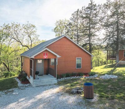 A small wooden cabin with a red exterior sits among green trees and grass. A gravel path leads to the front door, which has a small grill nearby. Another cabin is partially visible in the background.