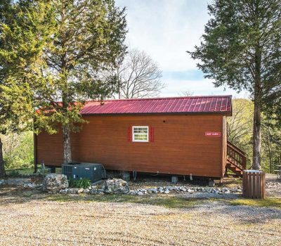 A small wooden cabin with a red metal roof is surrounded by trees. The cabin has a single window with white trim, a sign near the door, and a gravel driveway in front. Picnic tables and a trash bin are nearby.