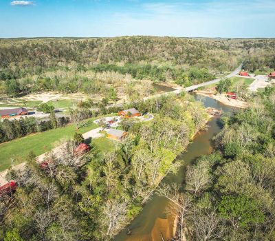 Aerial view of a river winding through a lush, green landscape with trees, scattered buildings, open fields, and a bridge crossing the river under a clear blue sky.