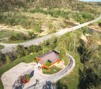 Aerial view of a red building with a curved driveway, surrounded by trees, near a road and river. A bridge crosses the river and leads into a forested, hilly landscape in the background.