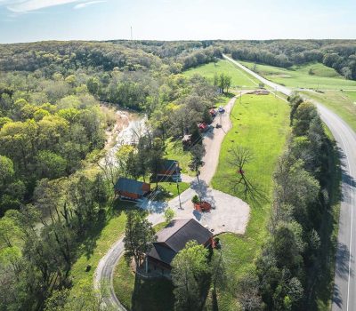 Aerial view of a rural landscape with winding roads, a few houses surrounded by trees, green grassy fields, and a small creek running through the wooded area. The sky is clear and sunny.