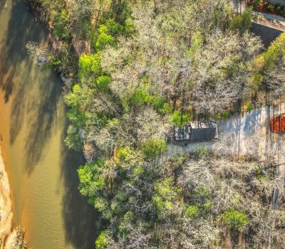 Aerial view of a river winding beside a wooded area with a house and gravel driveway partially visible among trees, some with green leaves and others bare.