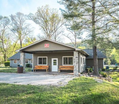 A single-story lodge with a covered porch, stone pillars, benches, and a red sign above the door sits among tall trees and greenery on a sunny day. A gravel driveway leads up to the building.