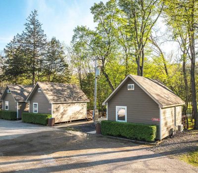 Three small brown cabins with peaked roofs are lined up beside a gravel road, surrounded by green trees and shrubs on a sunny day in a wooded area.