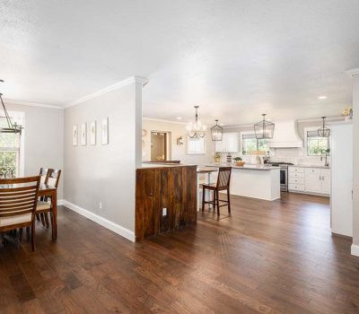 Open-concept dining and kitchen area with hardwood floors, a wooden dining table with six chairs, white cabinets, pendant lighting, and a partial dividing wall. Light streams in through multiple windows.