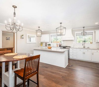Bright, modern kitchen with white cabinets, a large island, stainless steel appliances, wooden floors, and a small dining table with chairs. Three pendant lights hang from the ceiling, and windows let in natural light.