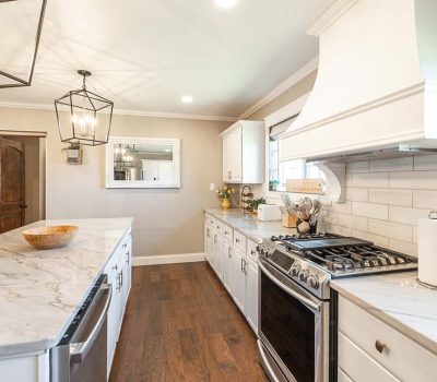 Bright, modern kitchen with white cabinets, marble countertops, a large island, stainless steel appliances, wooden floor, and neutral walls. Decorative lighting fixtures and wall accents add a cozy touch.