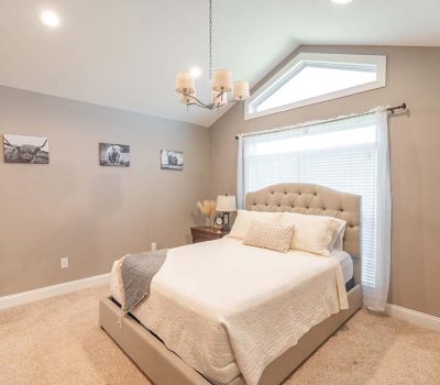 A bright bedroom with beige walls, carpeted floor, a large bed with a tufted headboard, white bedding, two nightstands, and three black-and-white photos on the wall. A window with blinds lets in natural light.