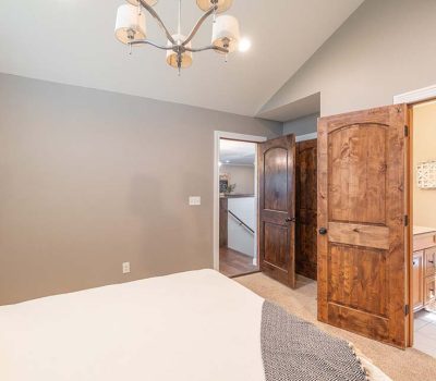 A cozy bedroom with gray walls, a beige bed, wooden double doors, and an adjacent bathroom visible with a toilet, vanity, and window. A modern ceiling light fixture hangs overhead.