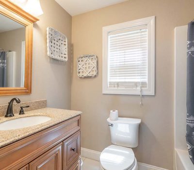 A small bathroom with a wooden vanity, granite countertop, and black faucet, a toilet with a roll of toilet paper, wall art, a window with blinds, and a bathtub with a gray patterned shower curtain.