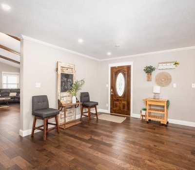 Bright entryway with wooden floor, a wooden front door with glass inset, two gray chairs by a decorative chalkboard, a small wooden cart with a plant, and a living area visible to the left.