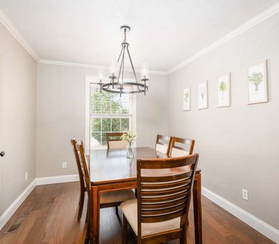 A bright dining room with a wooden table and six chairs, a modern black chandelier, light beige walls, wood flooring, framed botanical prints, and a window letting in natural light.