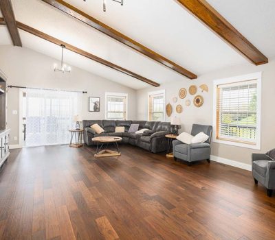 Spacious living room with dark wood floors, gray sectional sofa, accent chairs, wooden ceiling beams, wall art, large windows with blinds, and a media console. Natural light brightens the cozy, modern space.