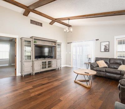Spacious living room with wooden floors, a sectional leather sofa, round coffee table, entertainment center with a large TV, exposed ceiling beams, and light coming through windows with white curtains.