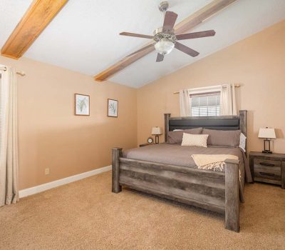 A cozy bedroom with beige walls, a large wood-framed bed with gray bedding, matching nightstands with lamps, two windows with blinds and cream curtains, a ceiling fan, and exposed wood beams on the ceiling.