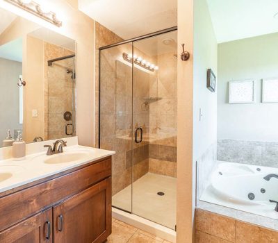 Modern bathroom with a glass-enclosed shower, double sink vanity with wood cabinetry, and a corner soaking tub with bronze fixtures. Neutral tiles and light walls create a bright, clean atmosphere.