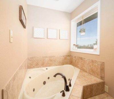 A corner bathtub with tan tile surround, dark fixtures, and a large window letting in natural light. Three framed wall art pieces and a hanging plant decorate the cozy bathroom space.